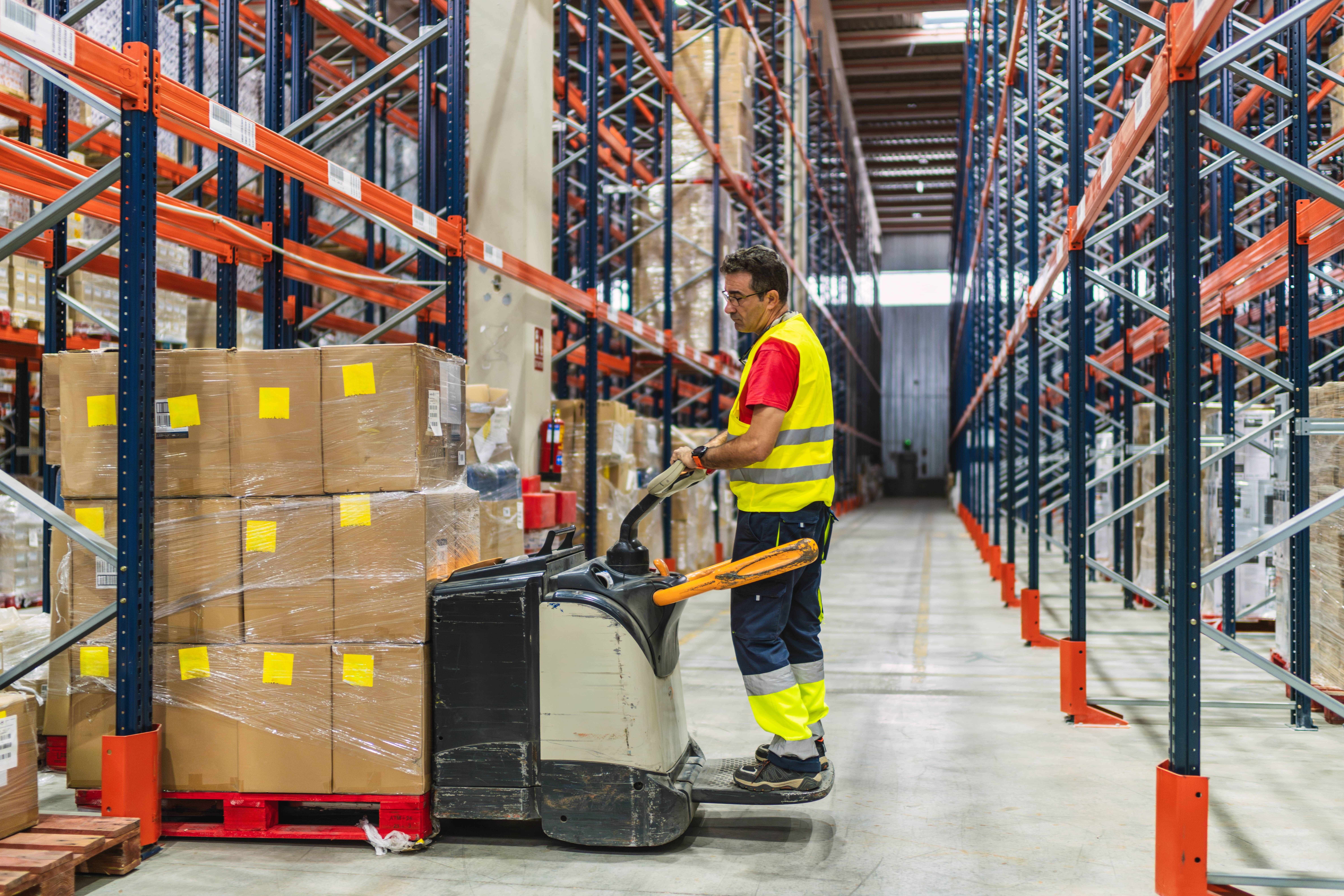 stock-photo-warehouse-worker-wearing-a-safety-vest-transporting-goods-on-a-pallet-jack-in-a-large-distribution-2544728975